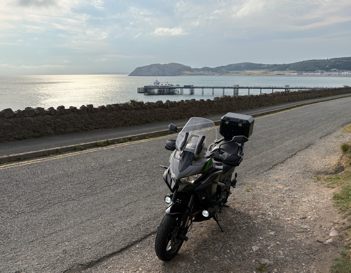 A Kawasaki Versys 1000 with a top box parked on a coastal road, with the pier and Great Orme headland at Llandudno across the bay
