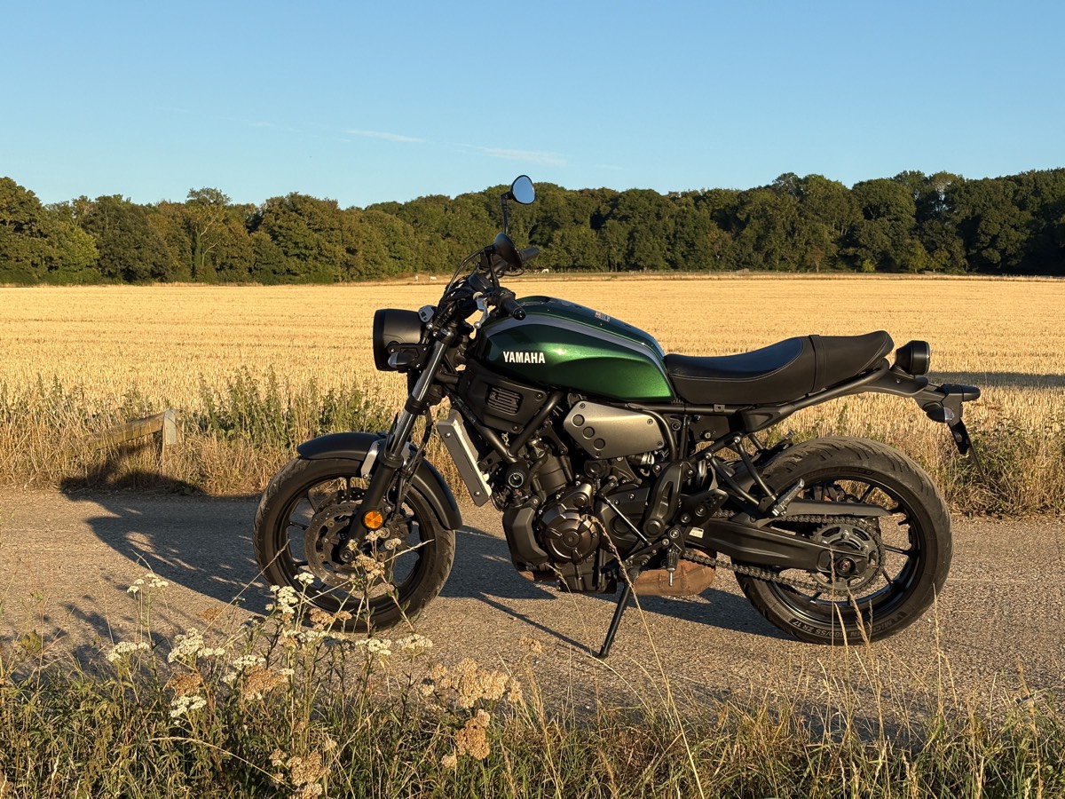 A green Yamaha XSR700 parked on a country lane beside a golden stubble field, with woodland behind