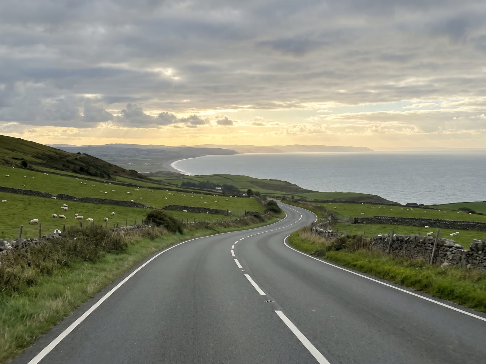 Near Aberaeron: a smooth two-lane road curving down a green Welsh hillside towards a distant sweep of grey-blue sea in Ceredigion, Wales