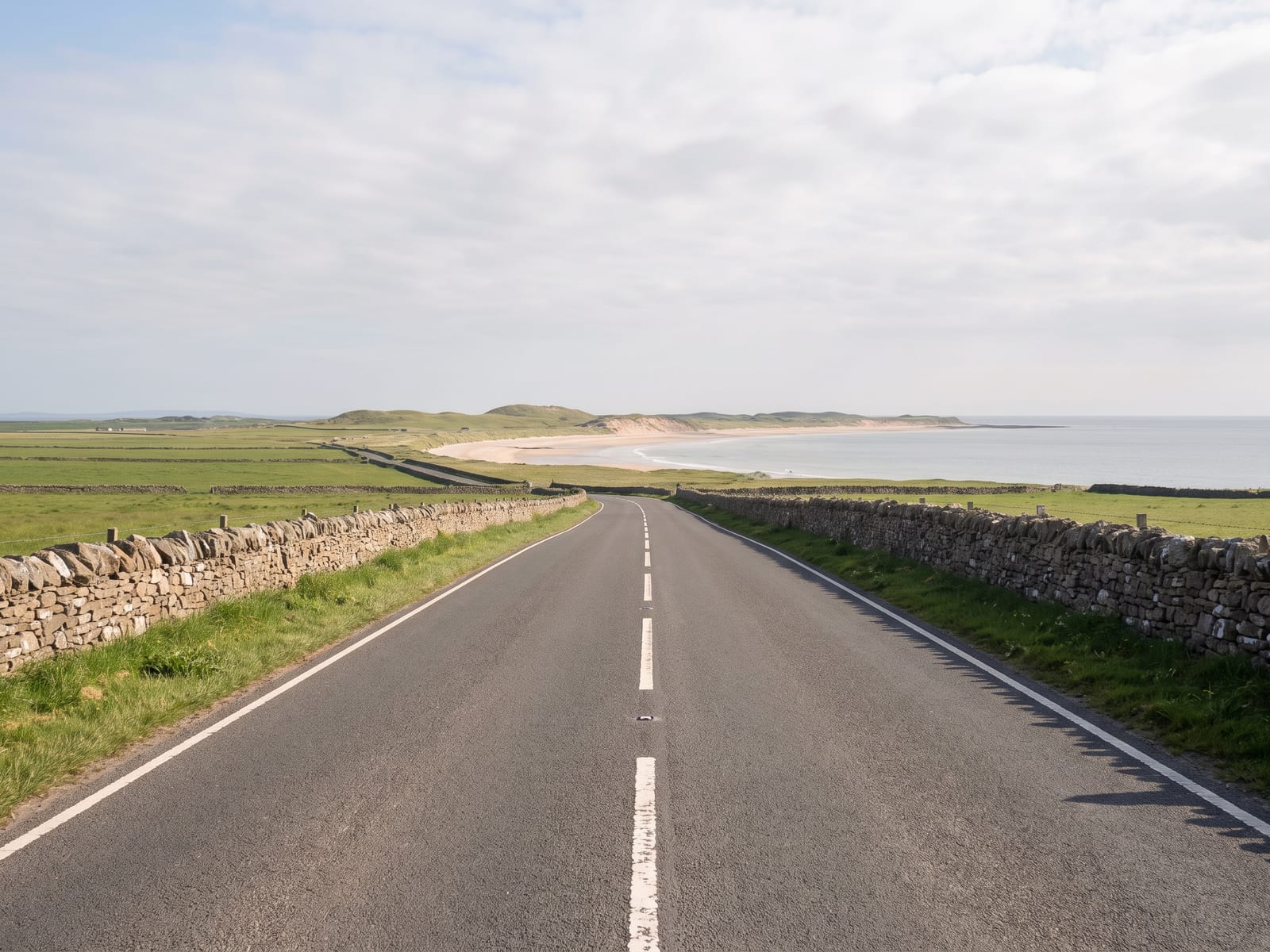 Near Alnwick: a smooth two-lane road running along flat farmland towards a pale sandy shoreline and calm grey-blue sea in Northumberland