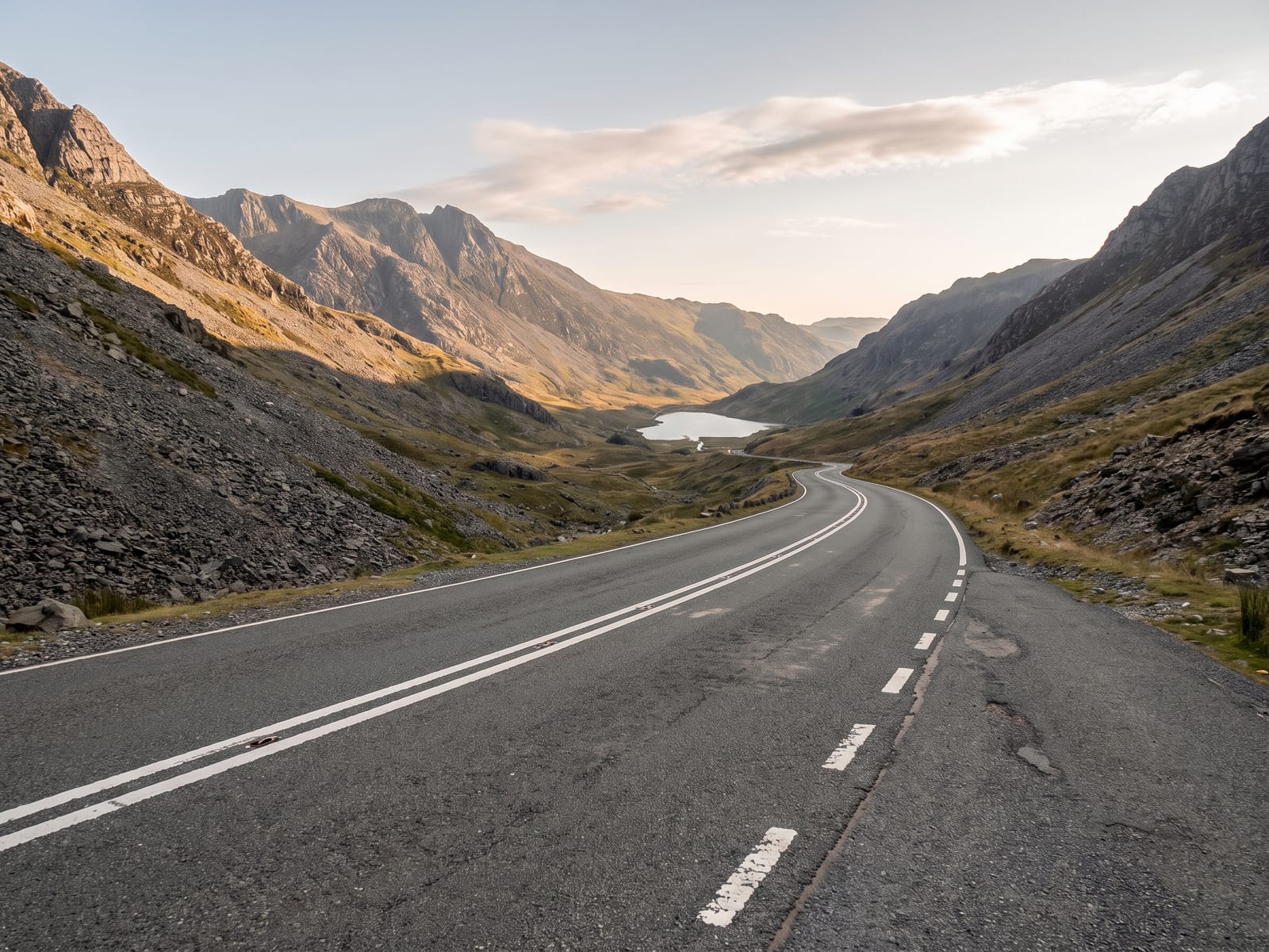 A wide sweeping tarmac road descending a mountain pass in Snowdonia, Wales, steep scree slopes and rugged grey peaks rising on either side with a small mountain lake glinting far below