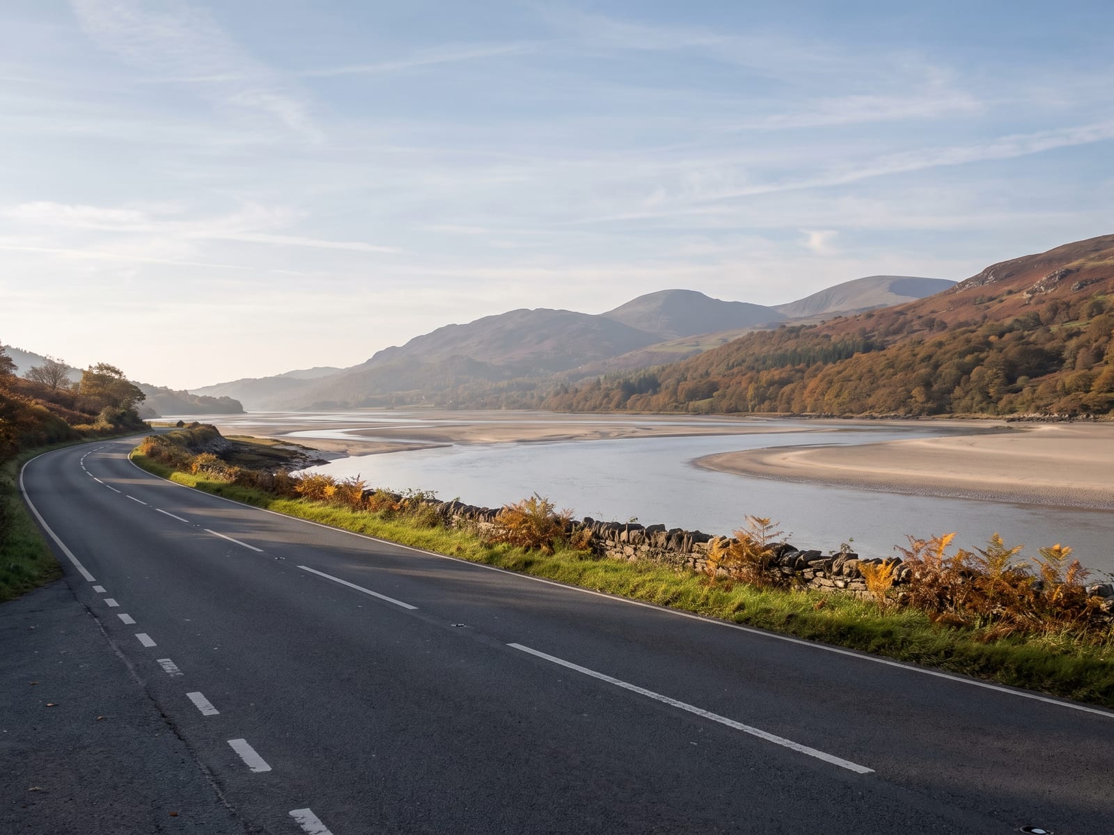 A smooth two-lane tarmac road running beside a wide tidal estuary on the Cambrian coast of Wales, pale sandbanks and calm silvery water to one side and wooded hillsides rising to rounded mountains on the other