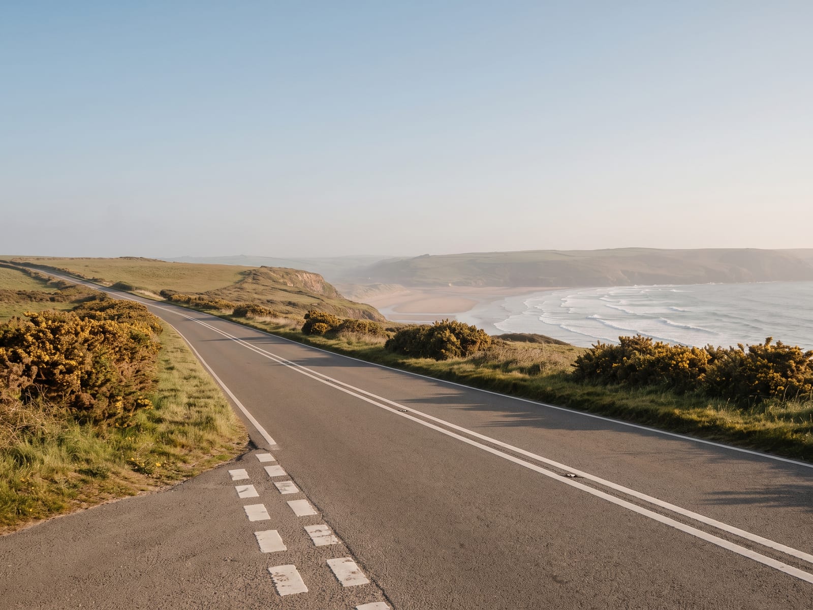 Near Barnstaple: a smooth two-lane road curving along a low clifftop above a wide sandy bay with rolling surf in North Devon
