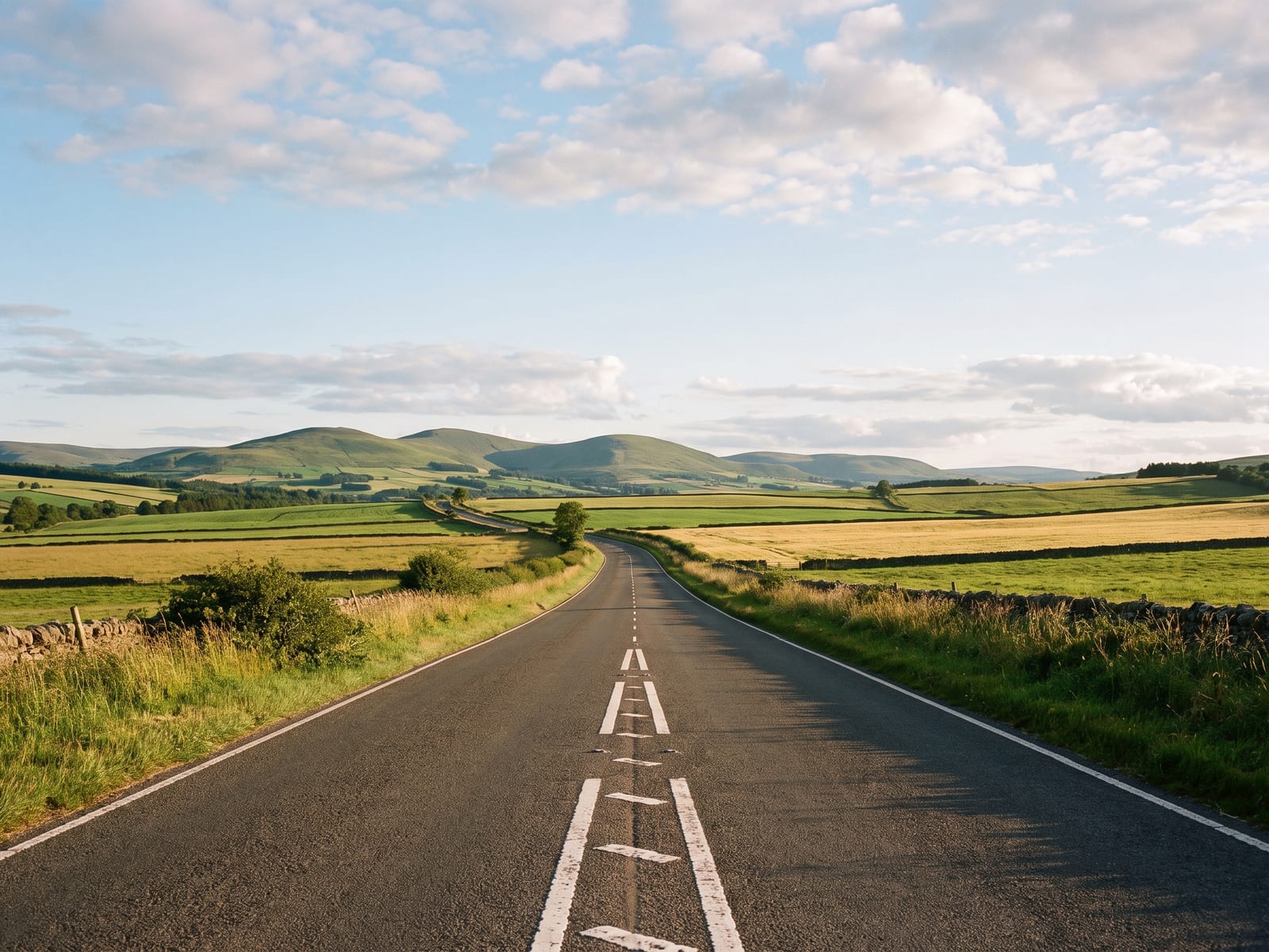 Near Berwick-upon-Tweed: a wide two-lane road crossing open rolling farmland with distant rounded green hills under a huge sky in the Scottish Borders, Scotland