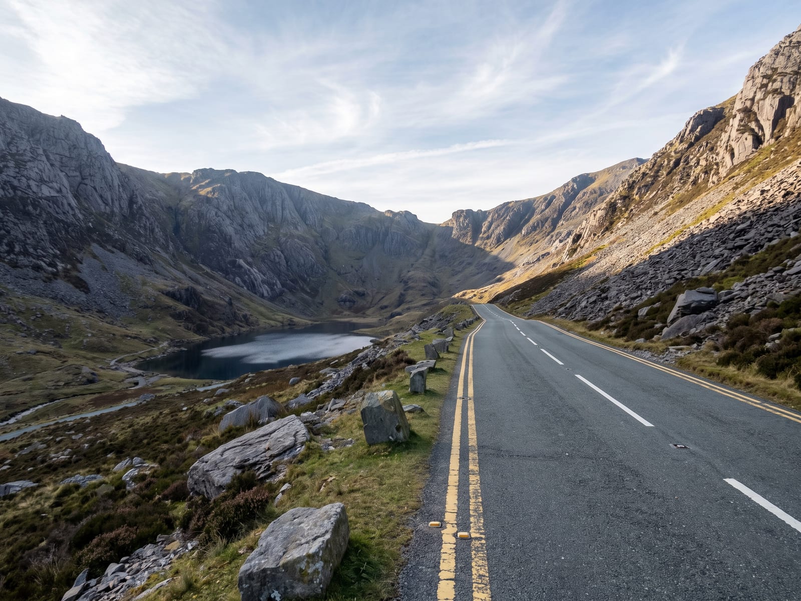 A tarmac mountain road climbing through a high glacial pass in Snowdonia, Wales, steep grey crags and scree slopes either side, a small dark lake in the valley below