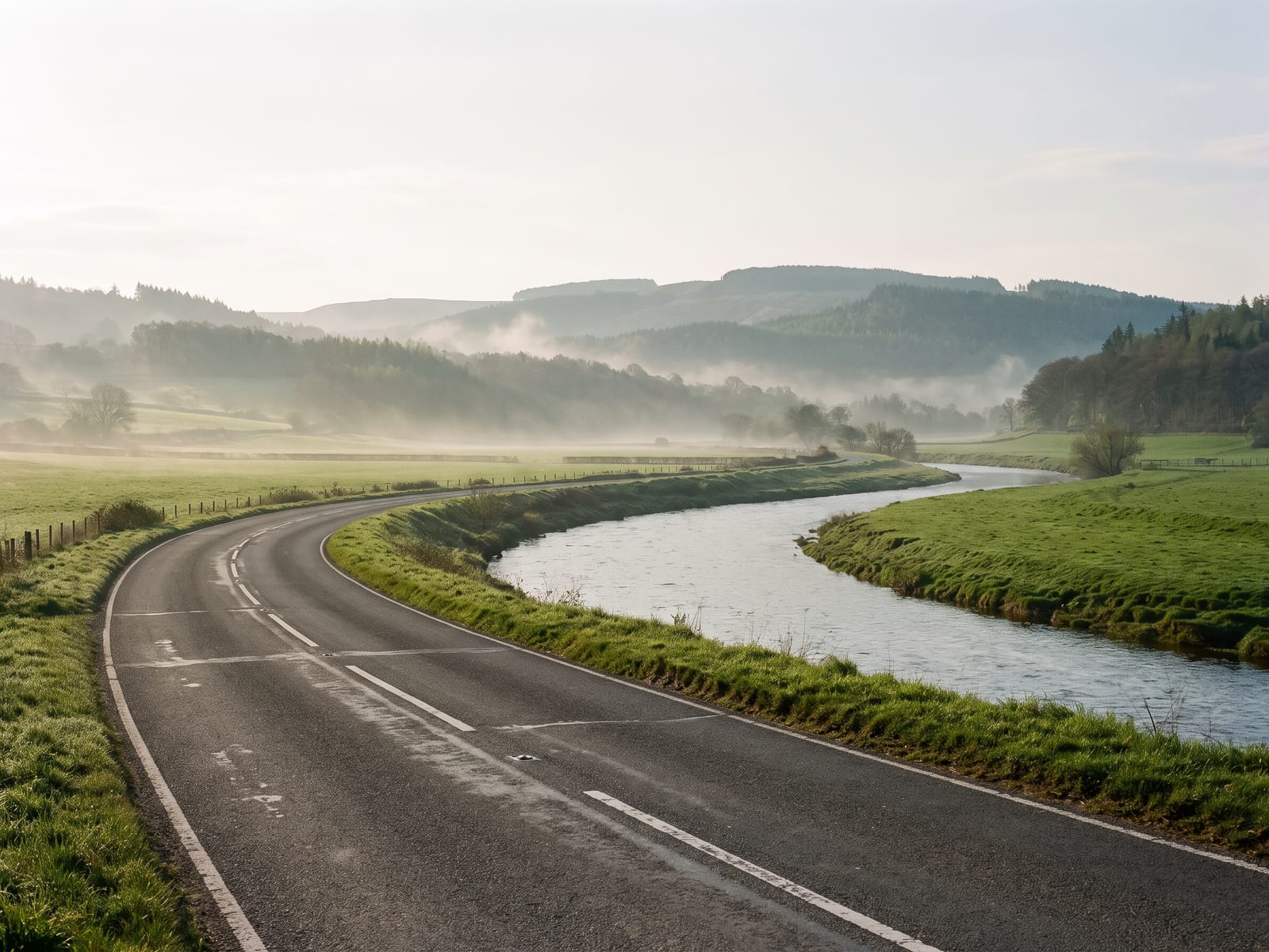 Near Builth Wells: a long open road running beside a wide meandering river through green farmland in Powys, Wales