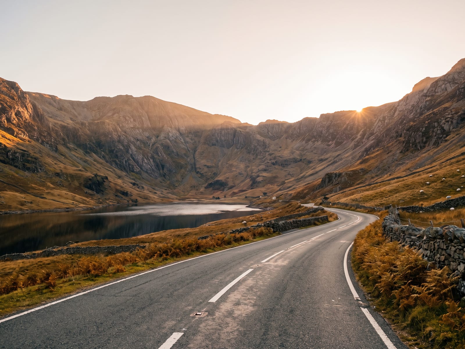 A tarmac road winding through a steep glacial valley in Snowdonia, Wales, with rocky mountain flanks catching golden light and a calm dark lake below the road