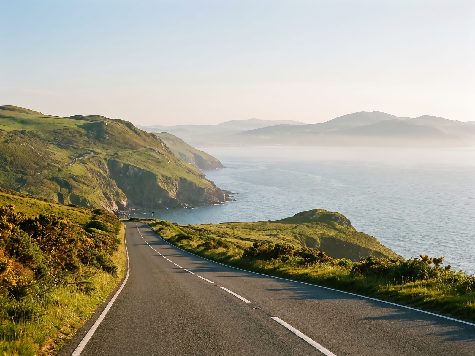 A tarmac coast road running along the edge of a rugged peninsula in North Wales, steep green headlands falling to a blue-grey sea and a distant line of hills across the bay