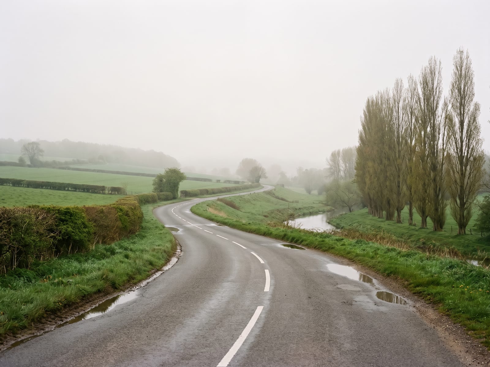 A gently rolling tarmac road in a quiet river valley in Norfolk, passing between hedged pasture and a shallow valley with a line of poplars