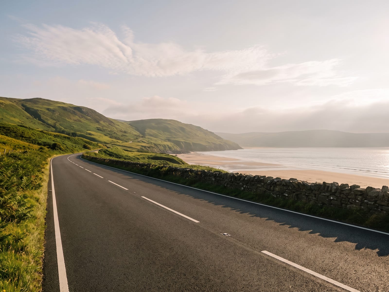 A smooth tarmac coast road running along the foot of green hills beside a wide sandy sea bay on the Cambrian coast, Wales, with a low stone wall on the seaward side and the sea glittering beyond