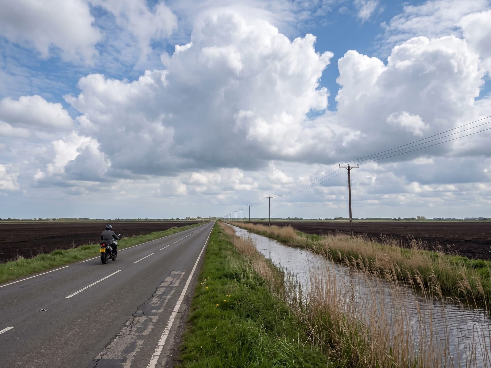 A dead-straight narrow tarmac road beside a wide fenland drain in Norfolk, black peat fields stretching flat to the horizon under an enormous sky of towering cumulus clouds