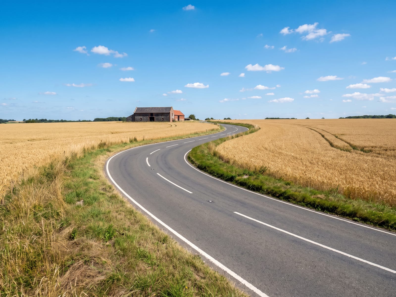 A smooth tarmac B road running straight then bending through open farmland in north-west Norfolk, wheat fields turning gold either side and a distant flint-walled farm building