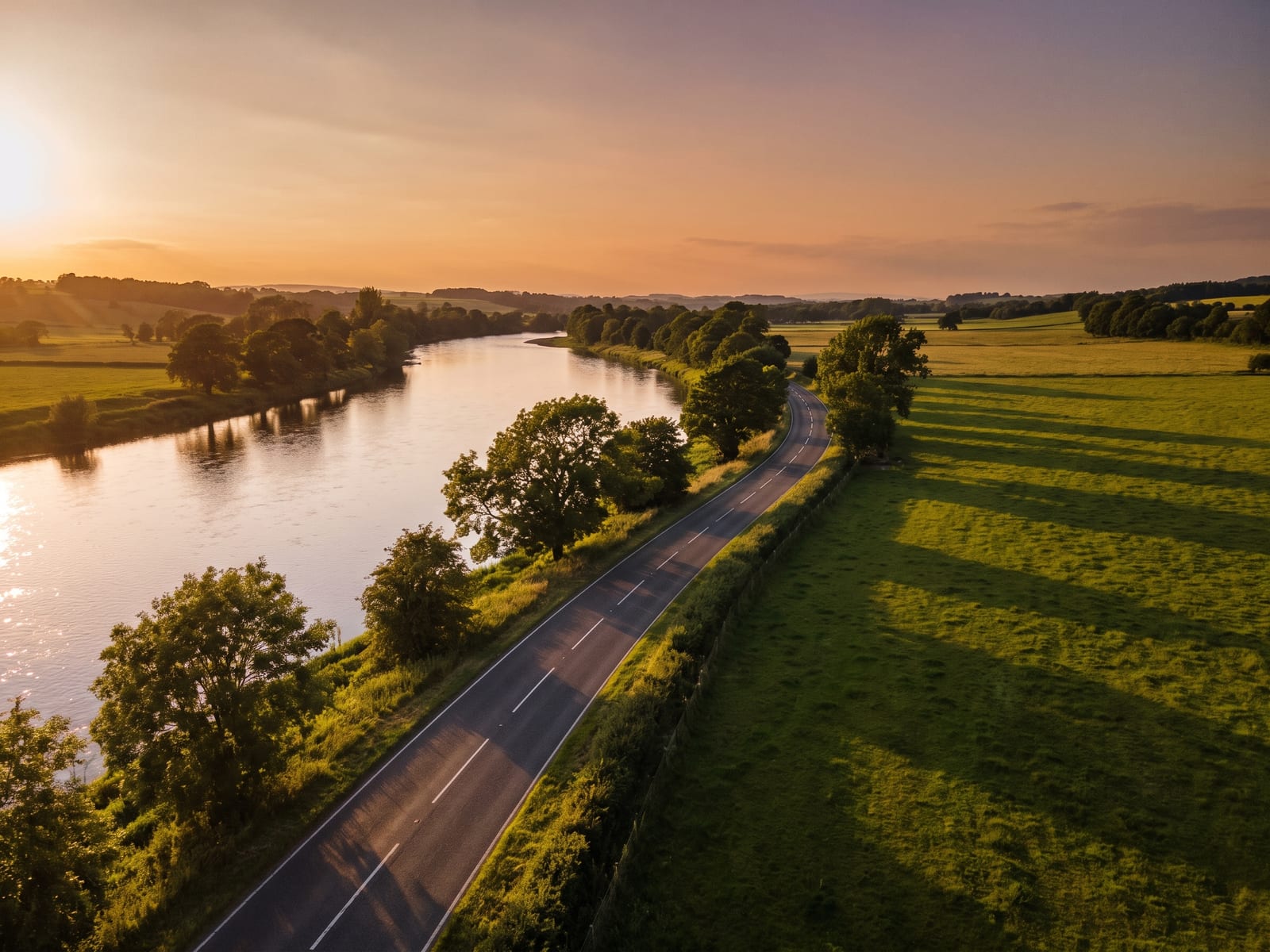 Near Kelso: a smooth country road beside a wide, slow river with tree-lined banks and gentle green fields in the Scottish Borders, Scotland