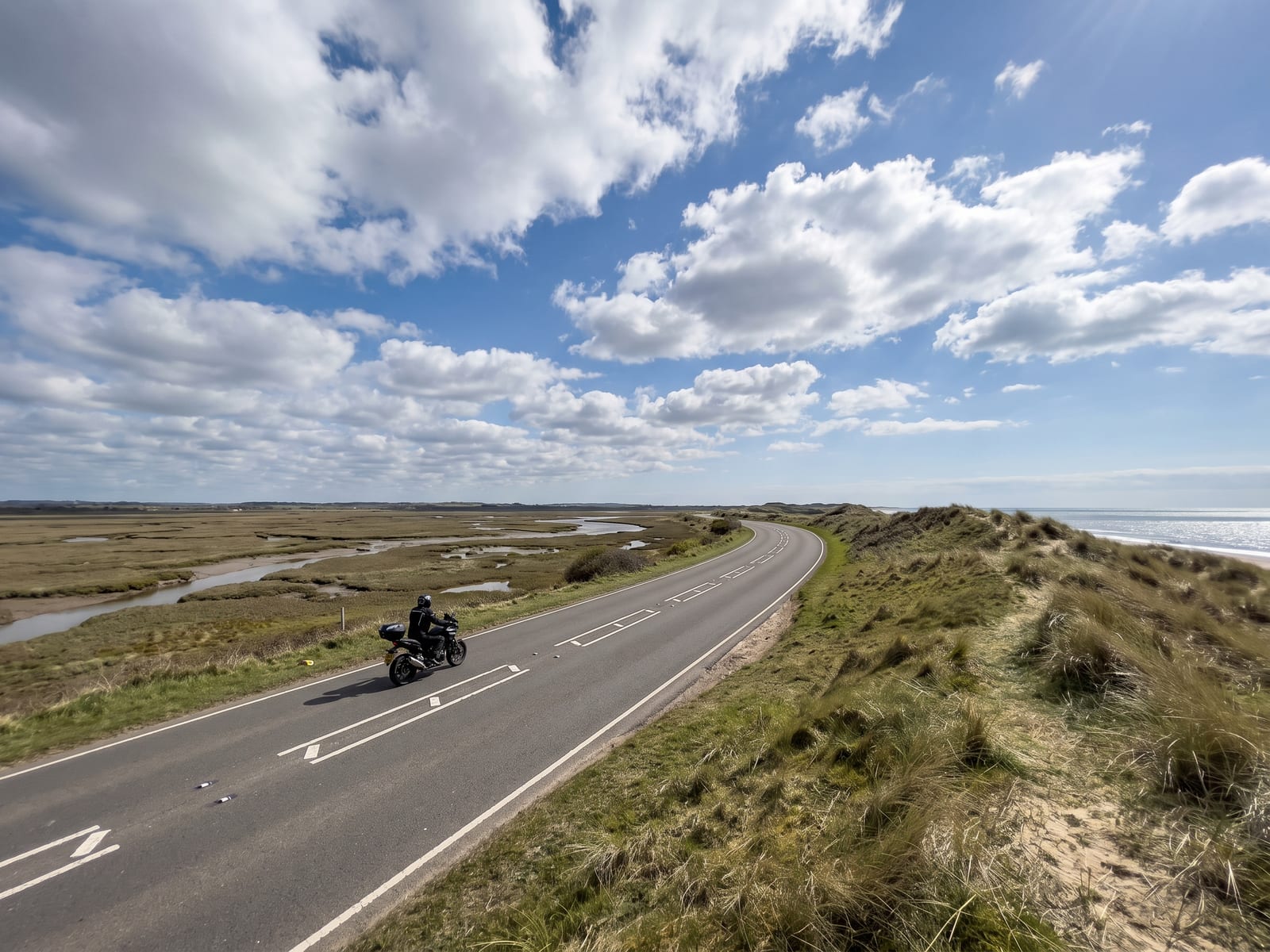 A smooth tarmac road on the Norfolk coast, running between salt marsh and a low dune ridge, with the sea a glinting strip beyond and a huge blue sky with scudding clouds