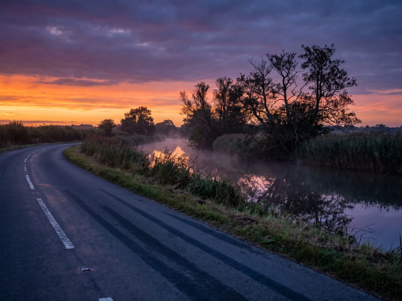 A narrow tarmac lane running beside a still river at dusk in Norfolk, with reed beds, alder trees and glassy water reflecting a deep orange and blue sky