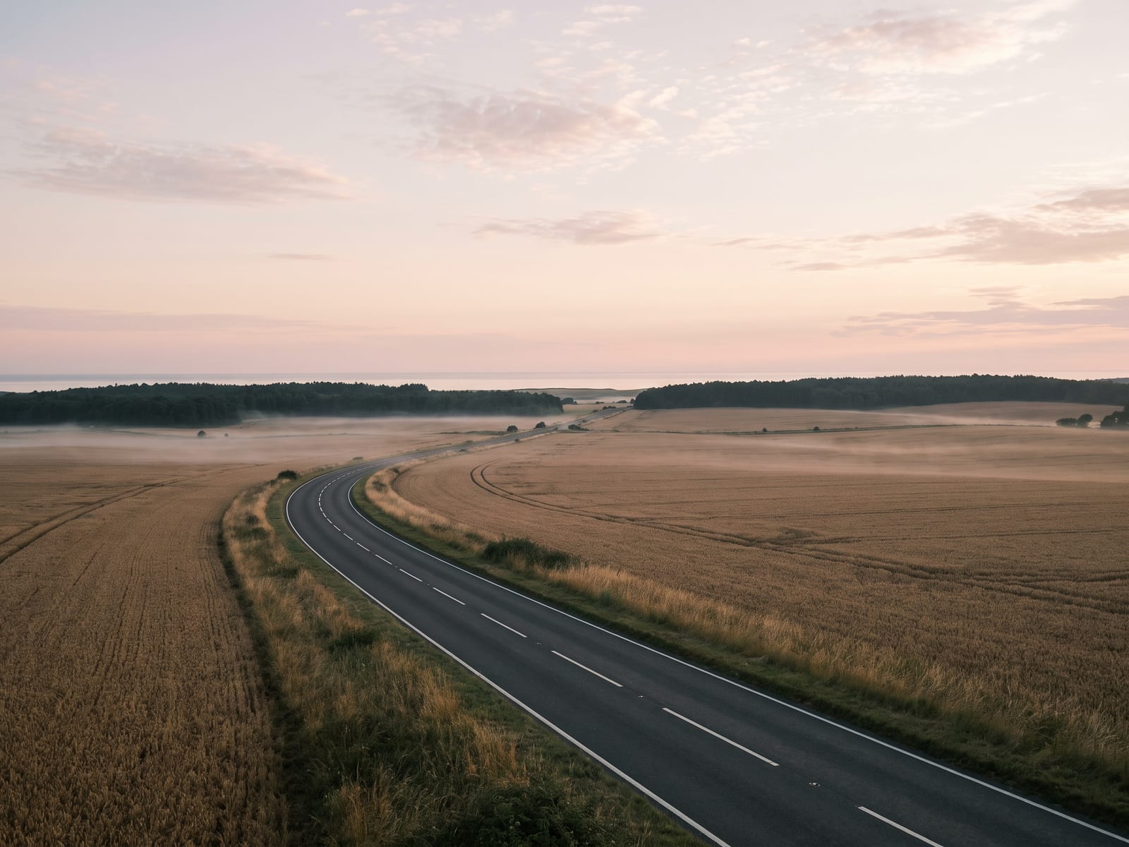 A sweeping tarmac A-road with fresh white edge lines crossing a broad open Norfolk landscape, wheat fields, a distant strip of dark forest and a pale sliver of sea on the far horizon