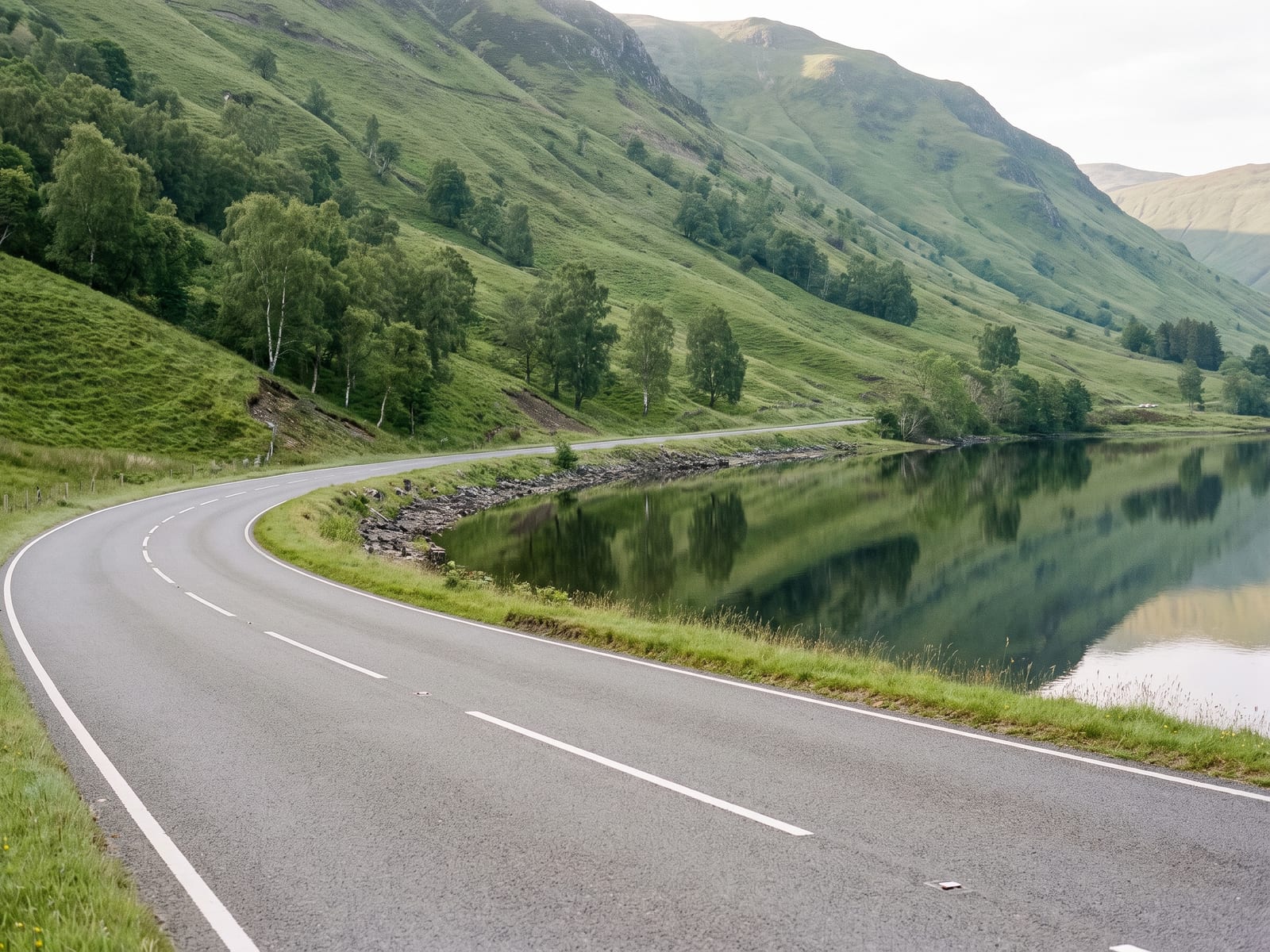 Near Peebles: a smooth two-lane road beside a long still loch with steep green hills rising behind in the Scottish Borders, Scotland